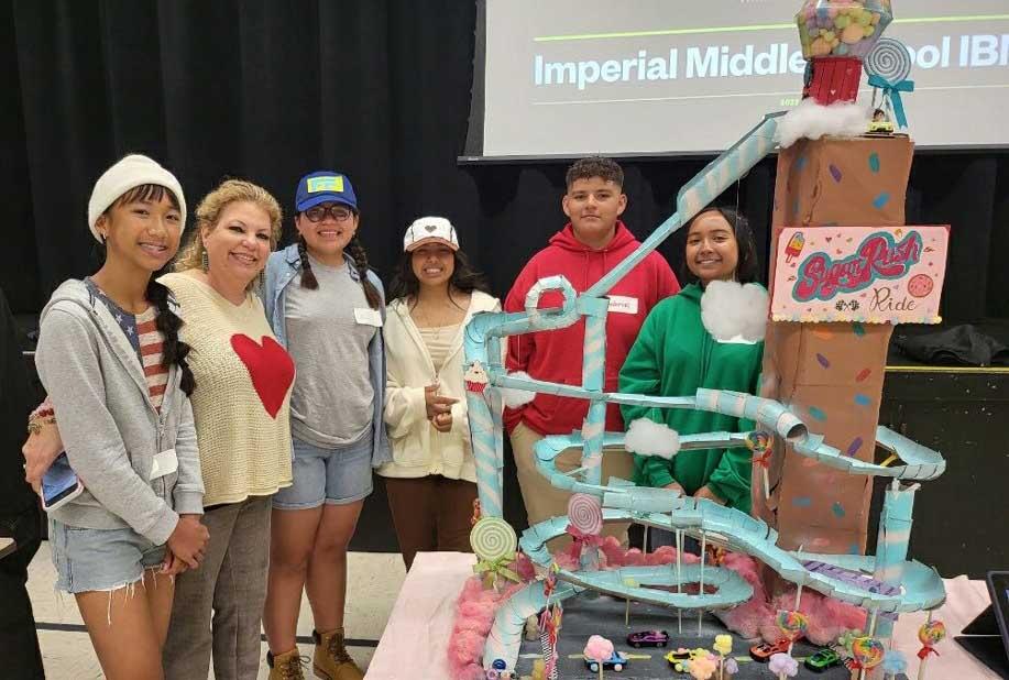 A group of cheerful students and a teacher standing proudly behind their creative project, a whimsical model of a roller coaster titled 'Sugar Rush Ride'. The students are dressed in casual attire, with some wearing hats and one in a festive red sweater. The project appears colorful and crafted with various materials, showcasing their ingenuity and teamwork. They are in a classroom with a presentation screen displaying 'Imperial Middle School | IB', indicating an educational setting focused on interactive and international learning.