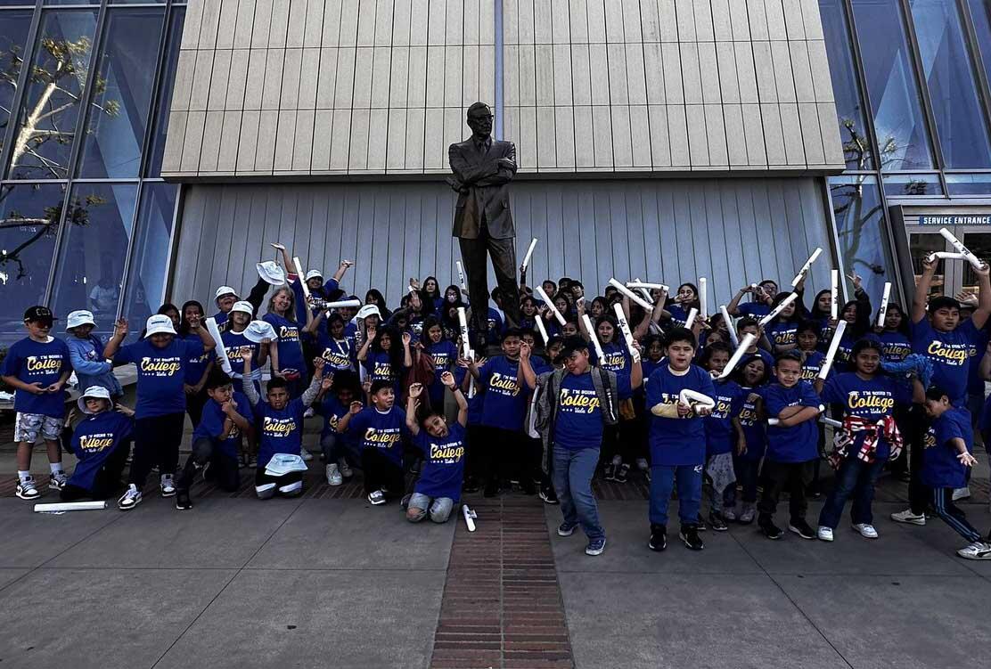 A lively group of children and adults gathered in front of a building, celebrating with arms raised and some holding rolled up posters. They're all wearing matching blue 'College' t-shirts. In the center of the photo, behind the group, stands a bronze statue of a man, which adds a sense of importance to the location. The setting suggests they are at a sporting event or participating in a team-related activity.